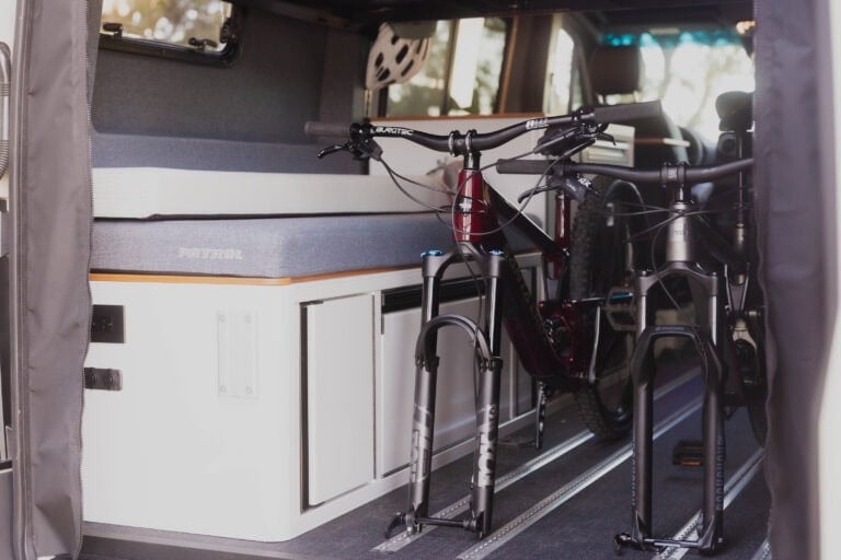 Two mountain bikes secured upright on an interior track floor system inside a Patrol Vans Sprinter conversion, alongside the bed platform and gear storage.