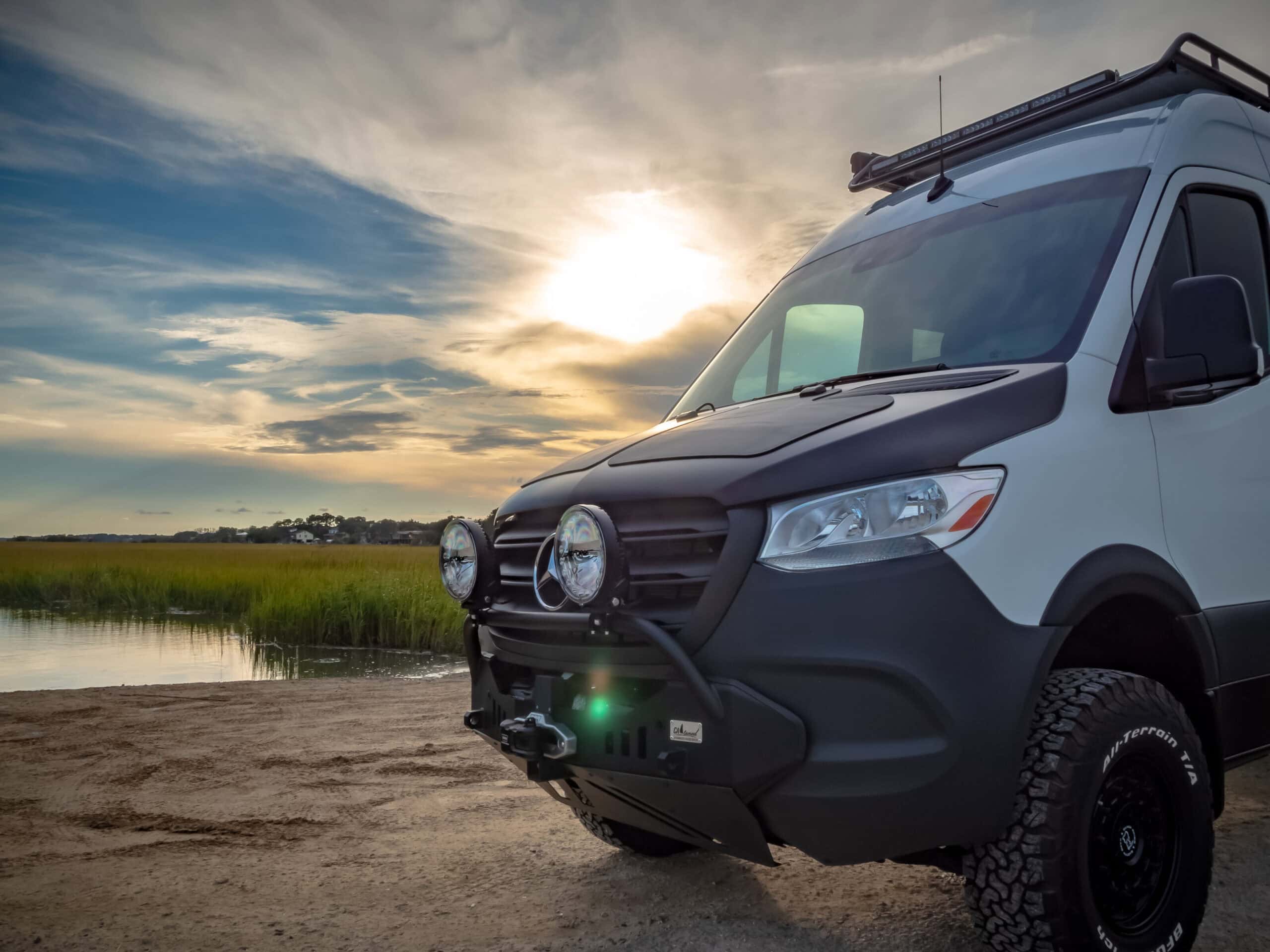 White Mercedes-Benz Sprinter conversion with an off-road front bumper, auxiliary driving lights, roof rack and BFGoodrich All-Terrain tires, parked on sand beside a Lowcountry marsh at sunset.