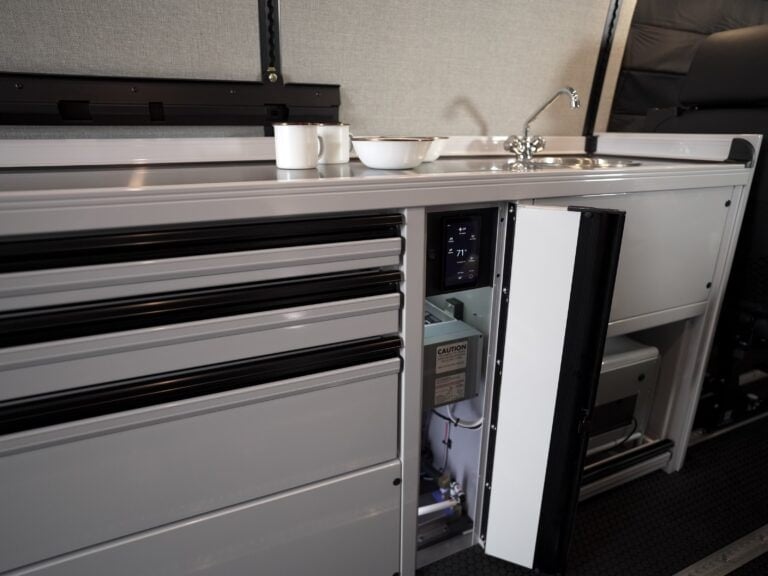 Open service bay in a Sprinter conversion galley showing the water heater unit, plumbing runs and a climate control touchscreen reading 71 degrees.