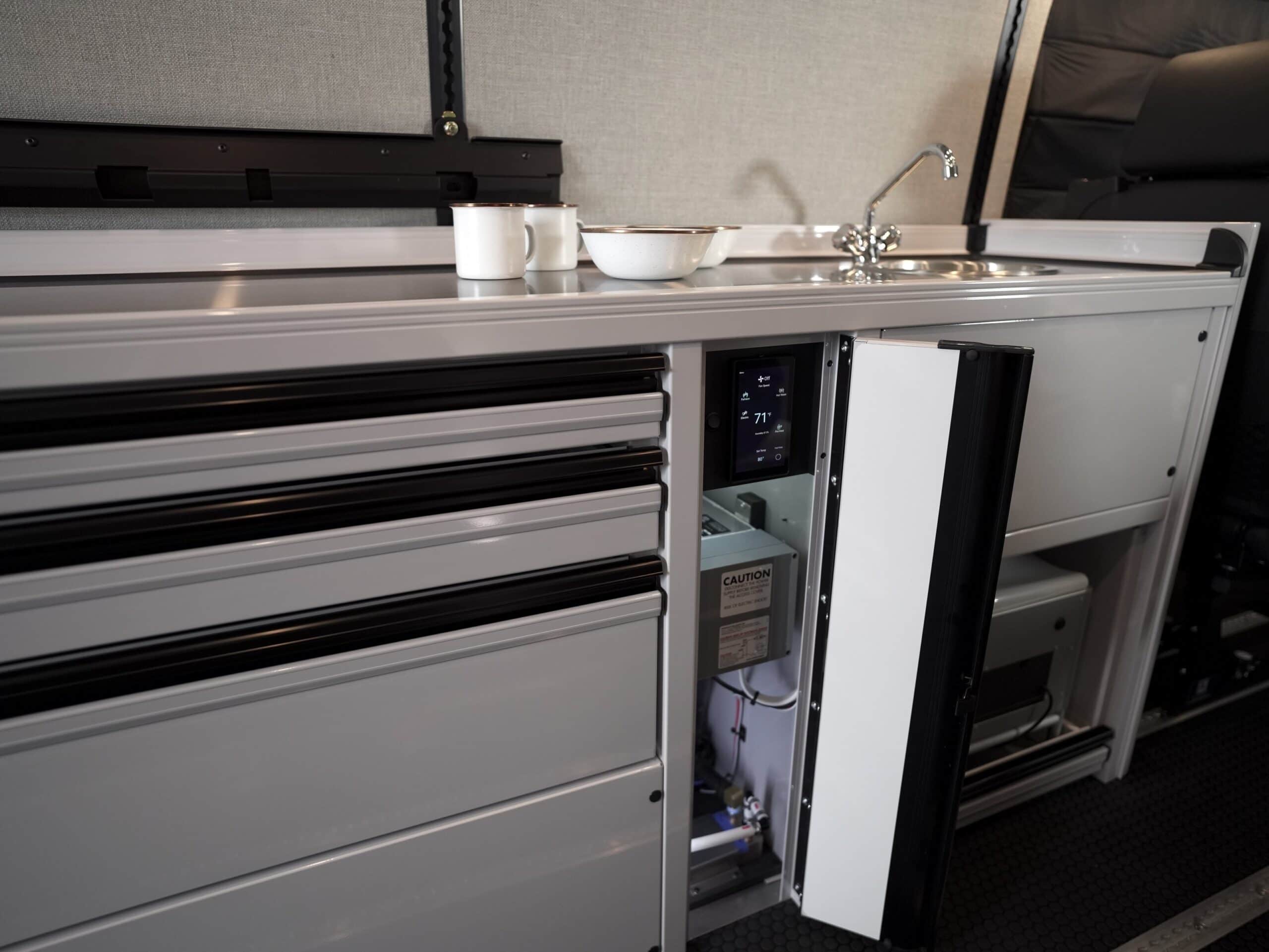 Open service bay in a Sprinter conversion galley showing the water heater unit, plumbing runs and a climate control touchscreen reading 71 degrees.