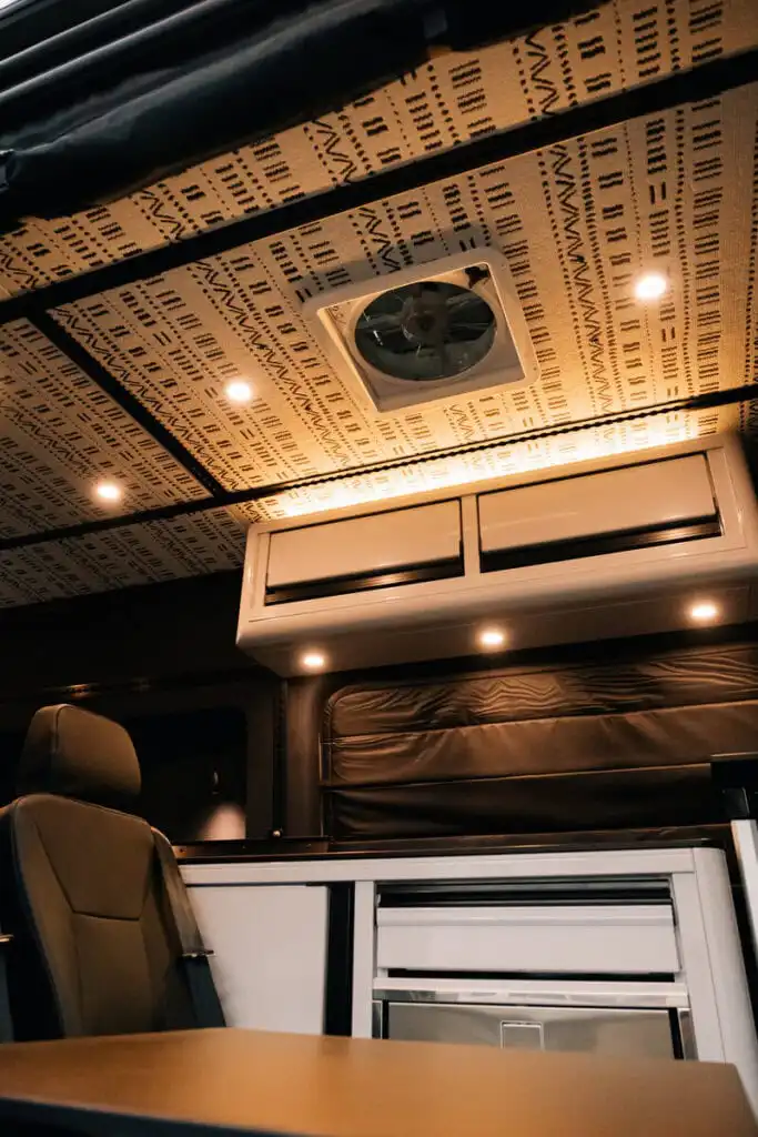 Interior of a camper van: patterned ceiling, roof vent, warm LED lights, and a beige seat nearby.