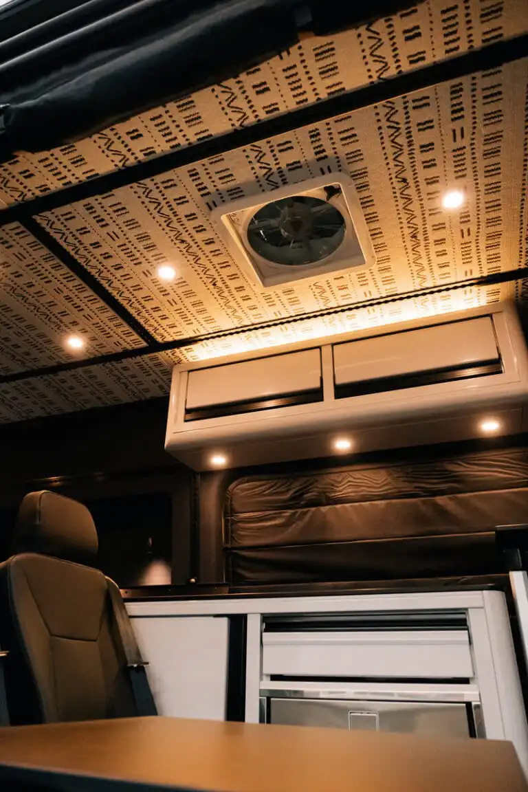 Interior of a camper van: patterned ceiling, roof vent, warm LED lights, and a beige seat nearby.