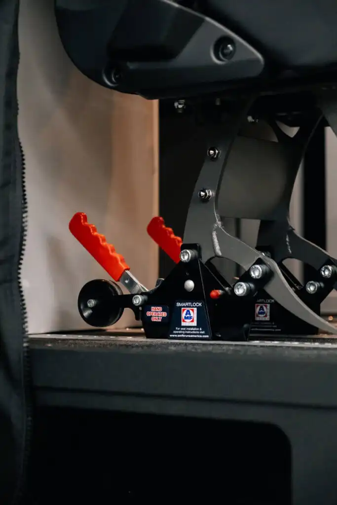 Close-up of a black metal locking mechanism with red hand grips and warning labels reading 'HAND OPERATED ONLY' and 'SMARTLOCK' on a chair or equipment base.