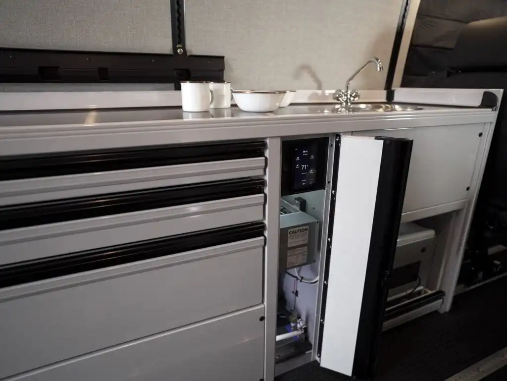 Metallic kitchenette in a vehicle with multiple drawers and a small open cabinet showing a control panel (71°) beside a sink and cups on the counter