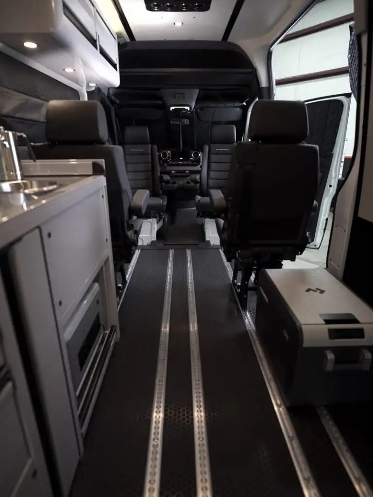 dsc06549 - Patrol Vans Interior of a modern passenger van: two front captain seats facing forward, central aisle, and equipment panels along the left side, with overhead storage and dim cabin lighting.