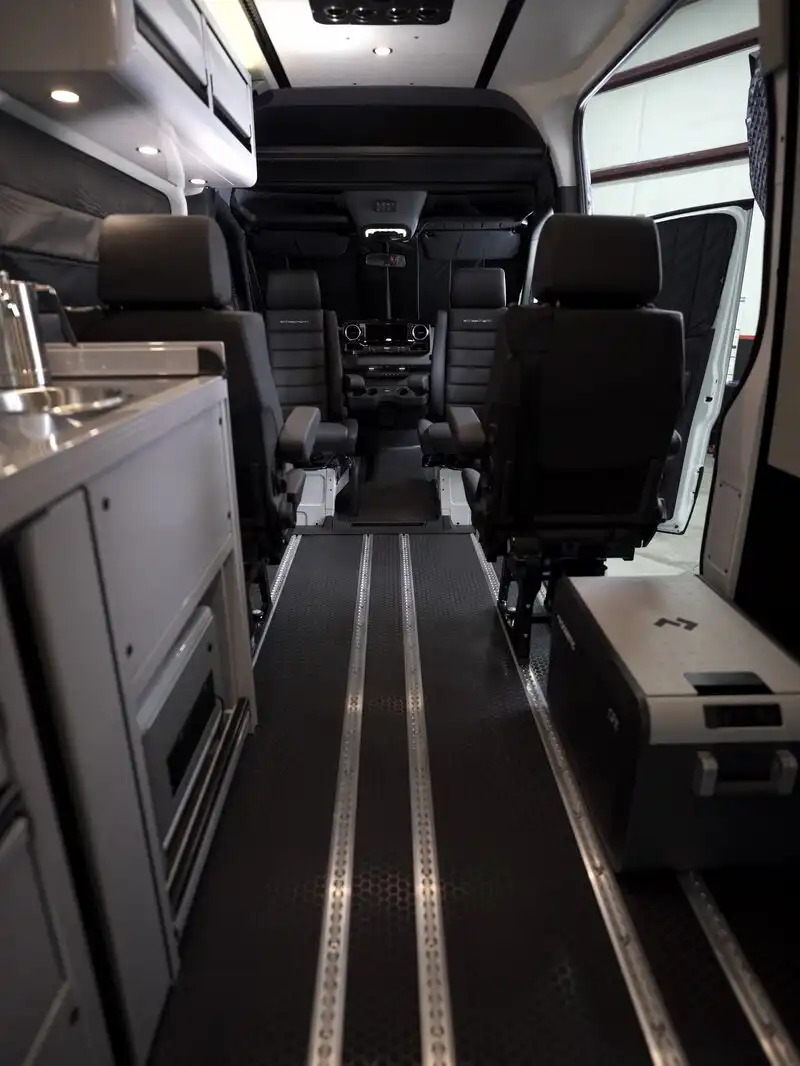 dsc06549 - Patrol Vans - Patrol Vans Interior of a modern passenger van: two front captain seats facing forward, central aisle, and equipment panels along the left side, with overhead storage and dim cabin lighting.