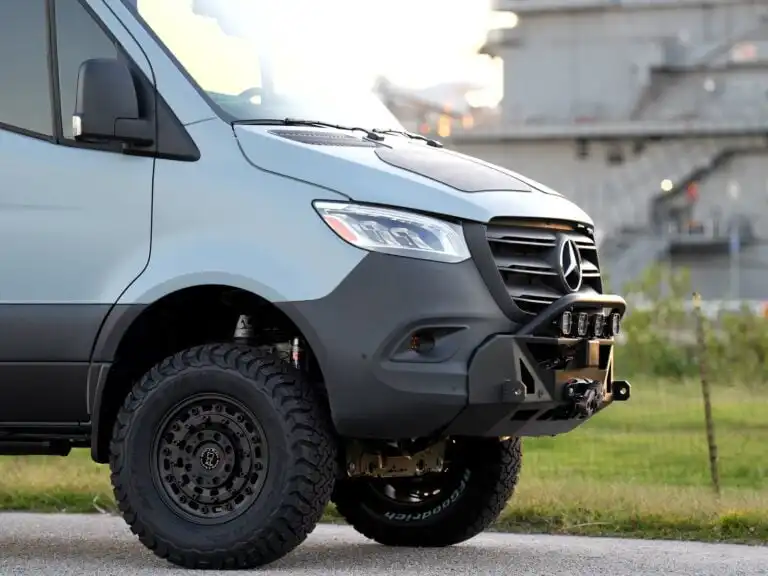 Front-left view of a light-blue Mercedes van with an aggressive off-road bumper and large tread tires, parked near an industrial backdrop.