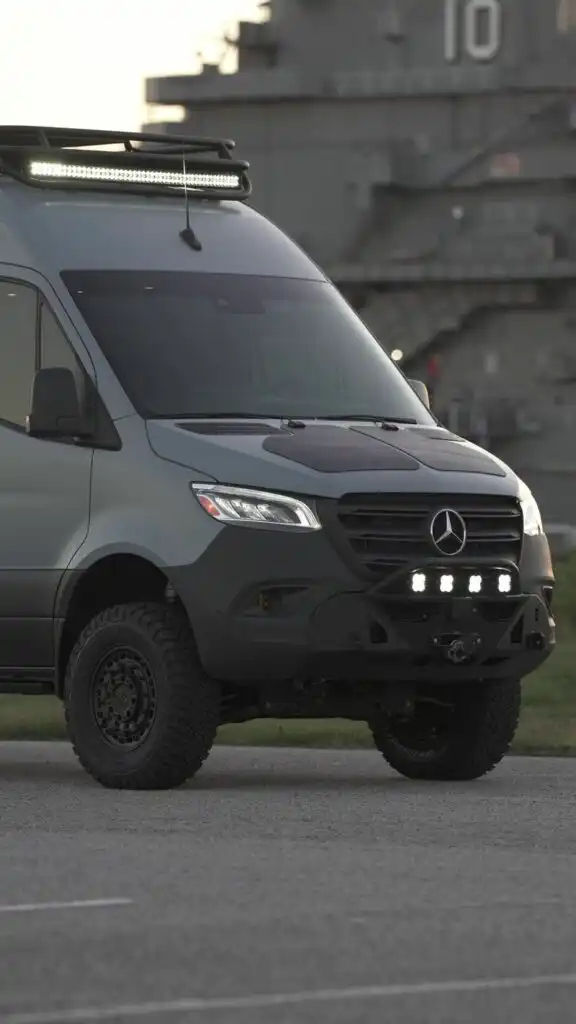 Best Custom Sprinter Van Builders in the US: How to Choose the Right One 2 Front three-quarter view of a modified gray Mercedes van with an LED roof light bar and rugged off-road bumper, parked on a concrete surface with a large naval ship in the background.