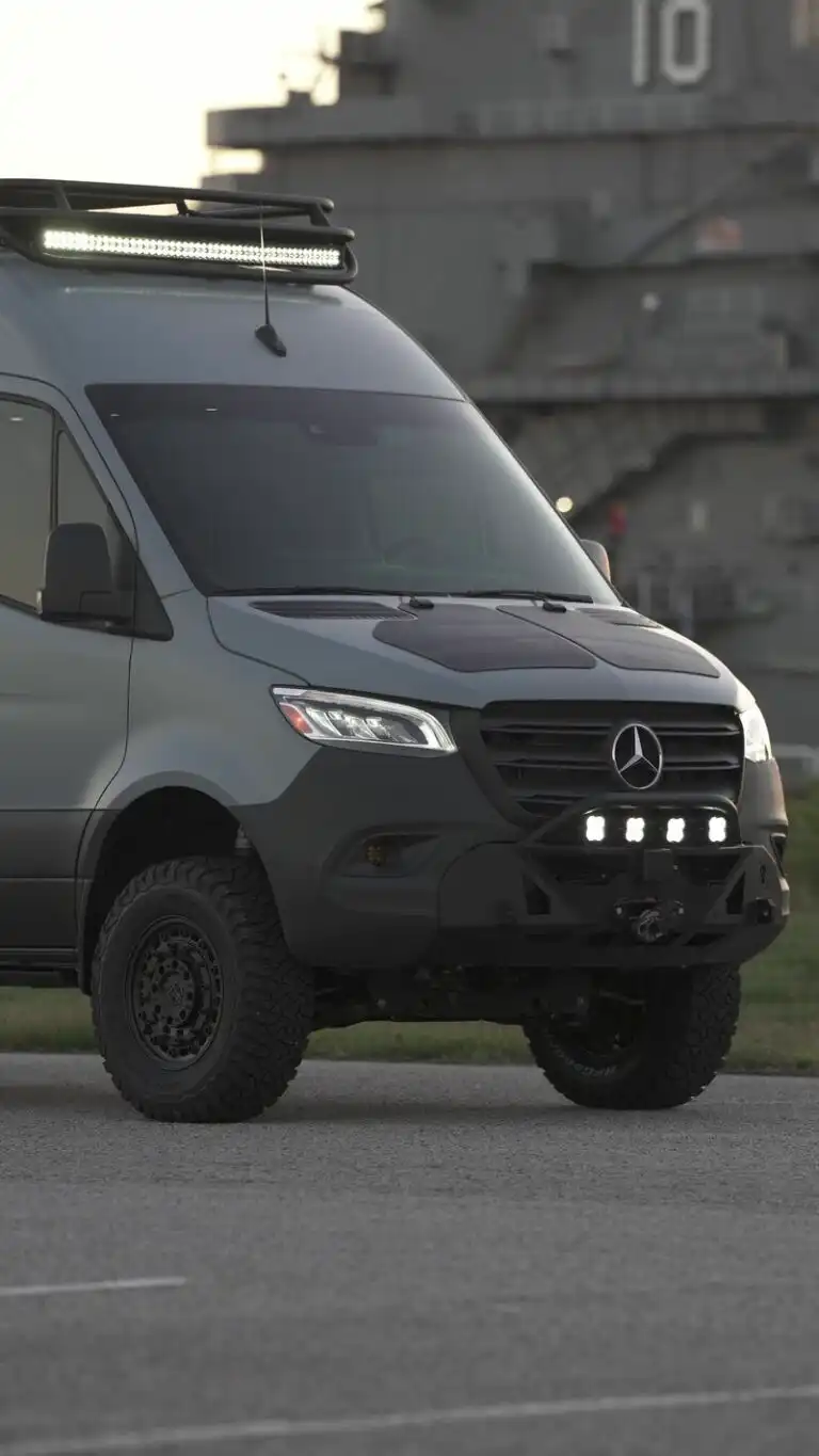 Front three-quarter view of a modified gray Mercedes van with an LED roof light bar and rugged off-road bumper, parked on a concrete surface with a large naval ship in the background.
