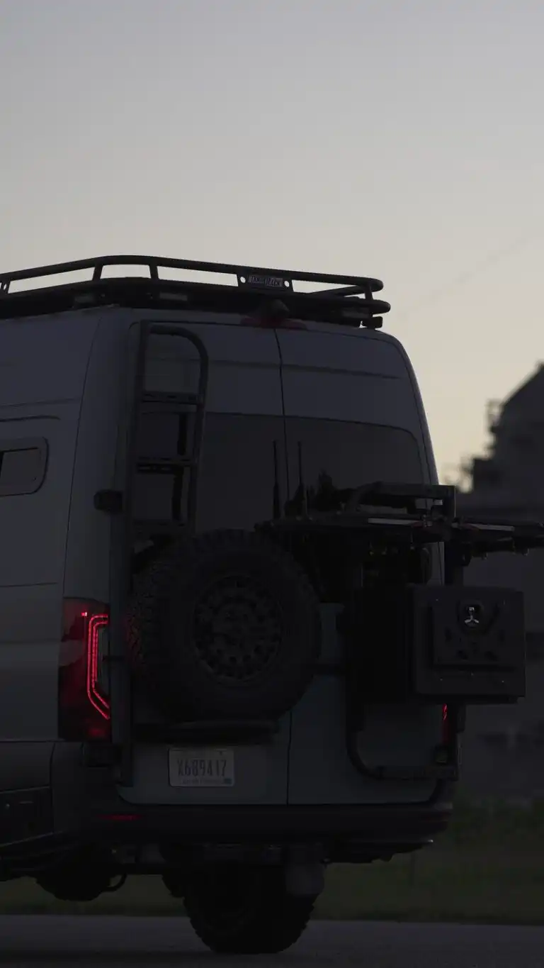 Rear view of a white van with a roof rack, spare tire, and bike rack at dusk, showing a South Carolina license plate.