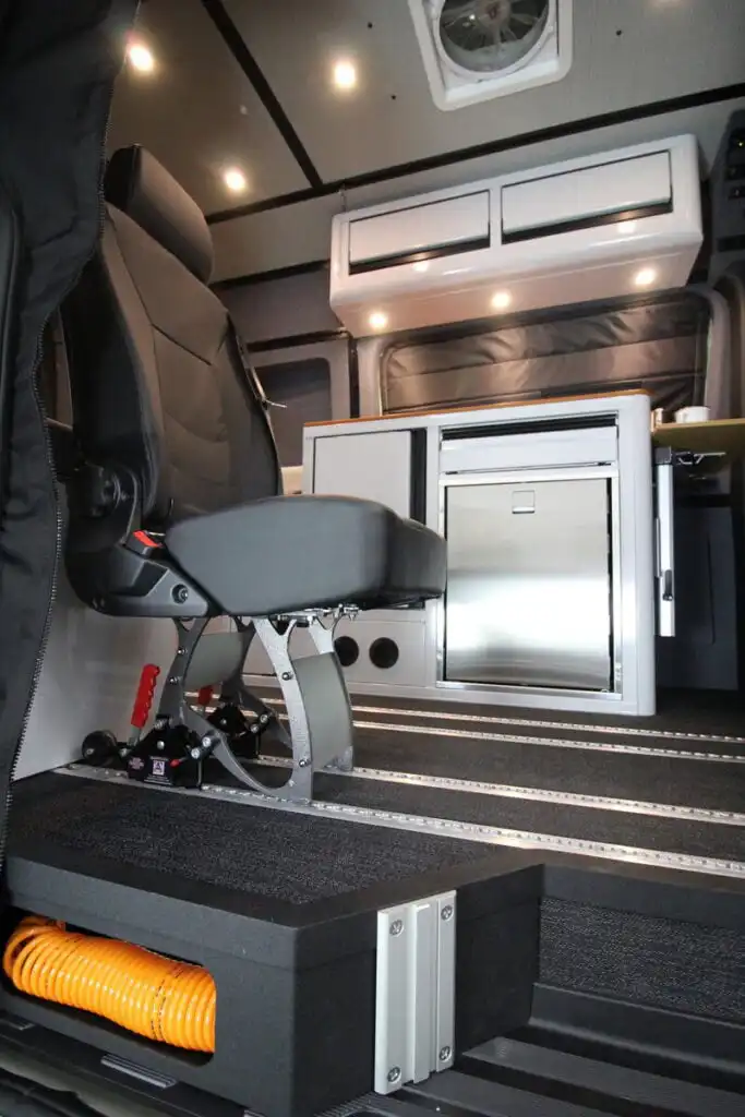 Interior of a mobile workspace showing a padded chair, stainless fridge, and overhead storage in a van or ambulance-type vehicle.
