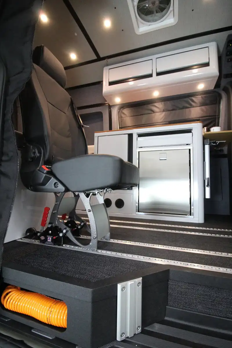 img_3662 - Patrol Vans - Patrol Vans Interior of a mobile workspace showing a padded chair, stainless fridge, and overhead storage in a van or ambulance-type vehicle.