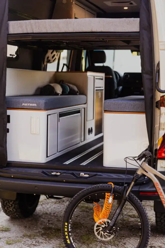 Camper van interior with a raised bed, sofa-style seating, and a compact kitchenette visible from the rear doorway; a mountain bike is locked outside.