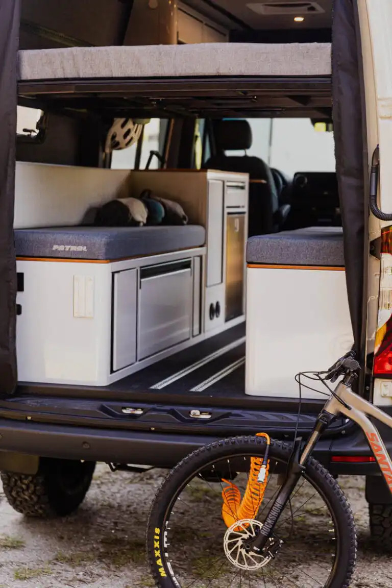 Camper van interior with a raised bed, sofa-style seating, and a compact kitchenette visible from the rear doorway; a mountain bike is locked outside.