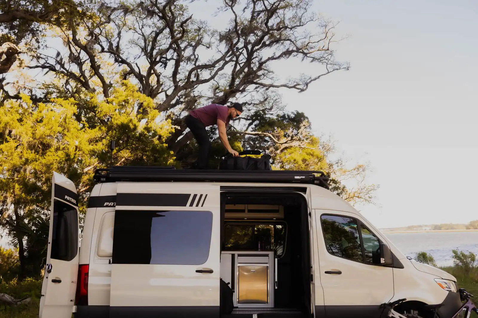 lowfeld_-0528 - Patrol Vans - Patrol Vans Man in a maroon shirt kneels on a camper van roof, securing gear on a roof rack beside a lake and trees on a sunny day