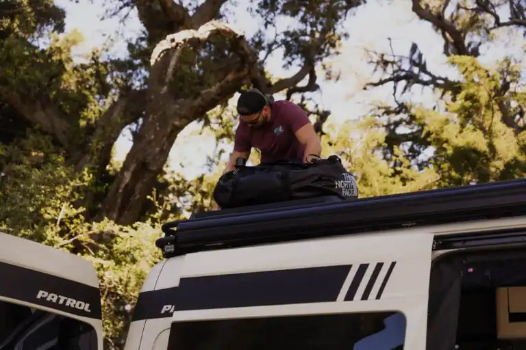 Man loading a large black duffel bag onto the roof rack of a white patrol van, with trees in the background.