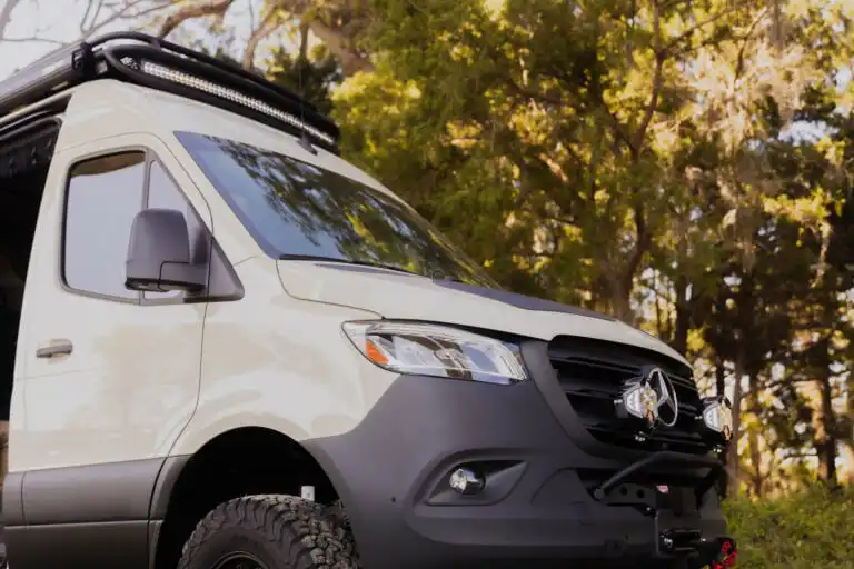 Front three-quarter view of a white off-road camper van with a roof rack and LED light bar among trees.