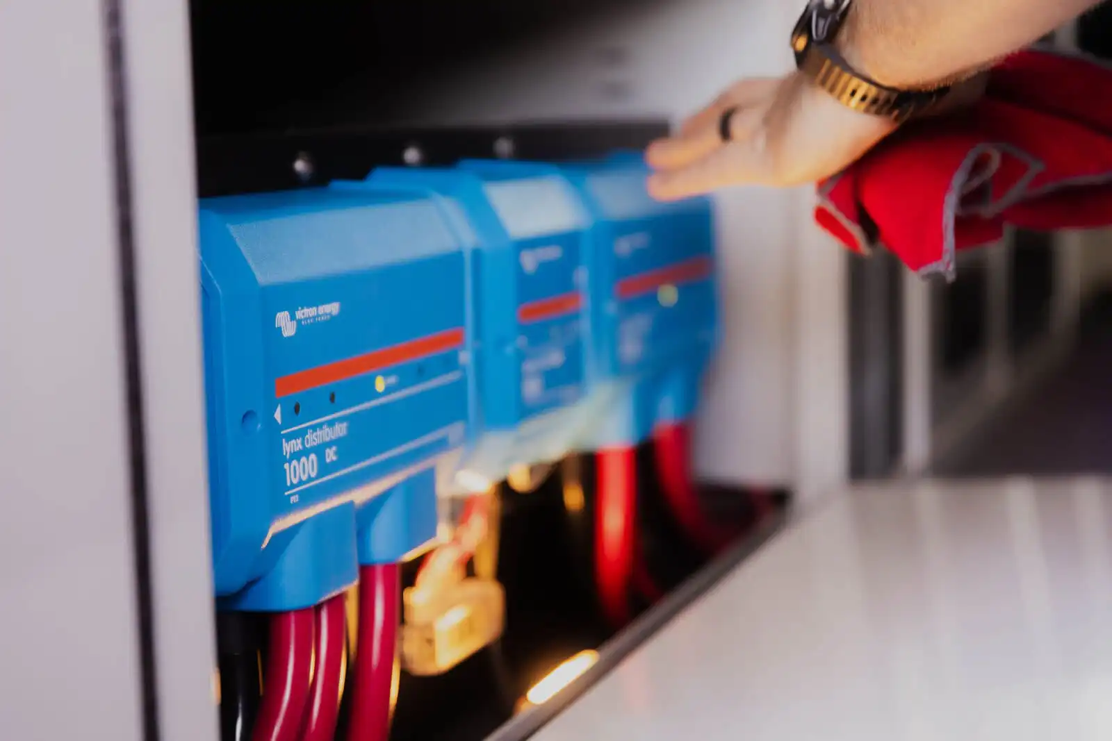 lowfeld_-0831 - Patrol Vans - Patrol Vans Blue Victron energy battery modules (1000 DC) with red cables in an electrical cabinet; a hand is nearby adjusting them.