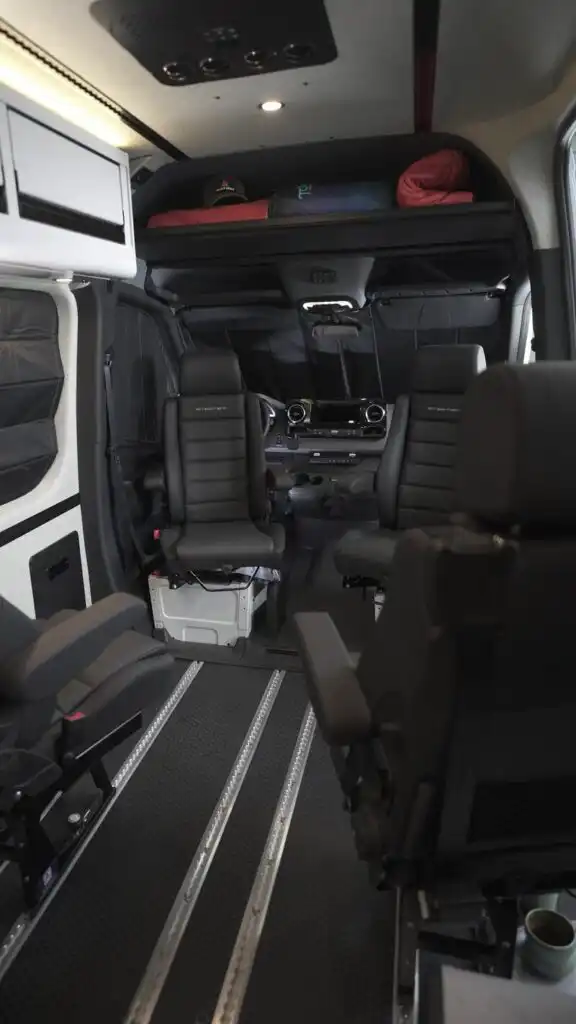 Interior of a van with two front captain chairs, overhead bunk/storage area with red blankets, and a metal floor track system behind the cockpit